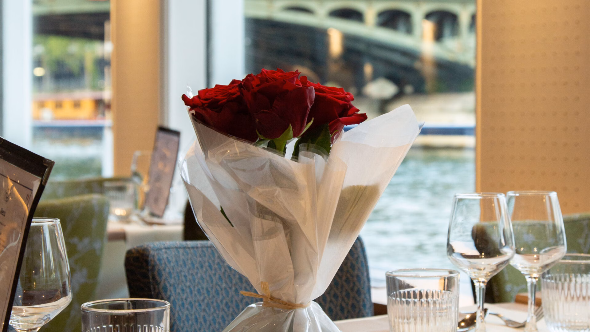 Table décorée avec un bouquet de roses rouges pour un dîner croisière romantique sur la Seine à Paris, offrant une vue sur un pont emblématique | Le Capitaine Fracasse